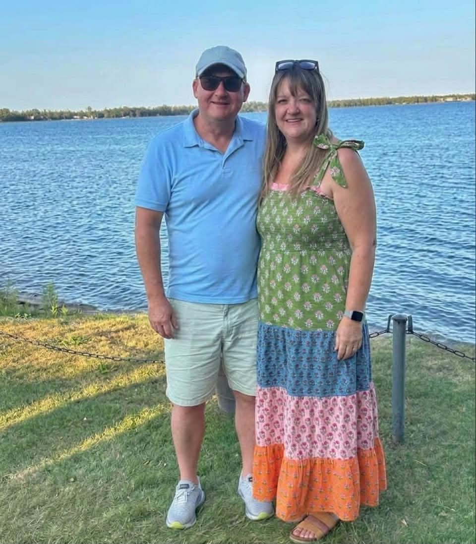 Adam and Beth standing together on a grassy shoreline, the bay behind them.